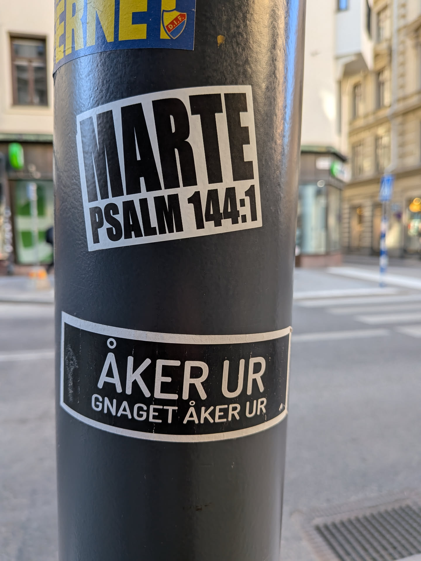 The sticker is rectangular with slightly rounded corners, featuring a stark black-on-white design. The word 'MARTE' is prominently displayed in large, bold, sans-serif capital letters across the top. Below it, in a smaller but still bold sans-serif font, is the text 'PSALM 144:1'. The sticker appears to be made of vinyl, showing minor signs of wear and adherence to the textured surface of the pole.