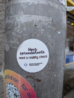 Street sticker A circular white sticker adhered to a weathered grey pole. The sticker features the main text "Your spreadsheets need a reality check" in a stylized font with a red and blue chromatic aberration or glitch effect, making the letters appear slightly offset. Below this text, there is a colorful, abstract logo composed of interconnected geometric shapes in shades of blue, purple, pink, and orange. To the right of the logo, the words "BALTIC SEA REGION YOUTH FORUM" are printed in a simple black sans-serif font.