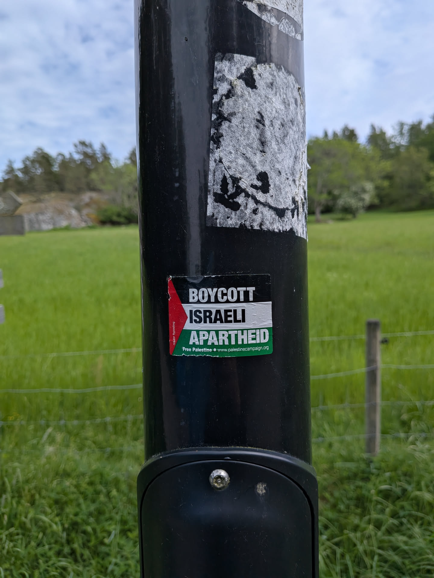 A rectangular sticker on a black pole featuring the Palestinian flag colors. It displays the text 'BOYCOTT ISRAELI APARTHEID' in bold white letters over black, white, and green stripes, with a red triangle on the left.