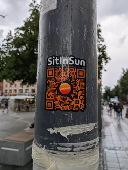 Street sticker Die-cut black sticker on a pole. Features white 'SitInSun' text, Swedish subtext, and a large orange QR code with a central orange gradient logo.