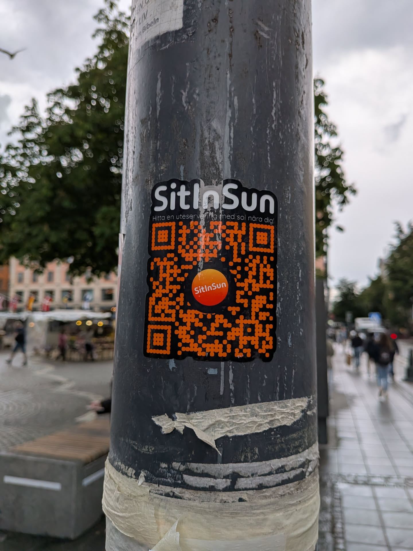 Die-cut black sticker on a pole. Features white 'SitInSun' text, Swedish subtext, and a large orange QR code with a central orange gradient logo.