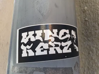 Black and white sticker with the text "KING KERZ" in a stylized graffiti font. There is also a black and white portrait of a person next to the text. The sticker is affixed to a gray metal surface.