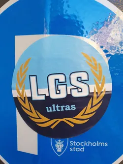 Round sticker with the text LGS ultras in the center, surrounded by a gold laurel wreath. The background is light blue and dark blue. 