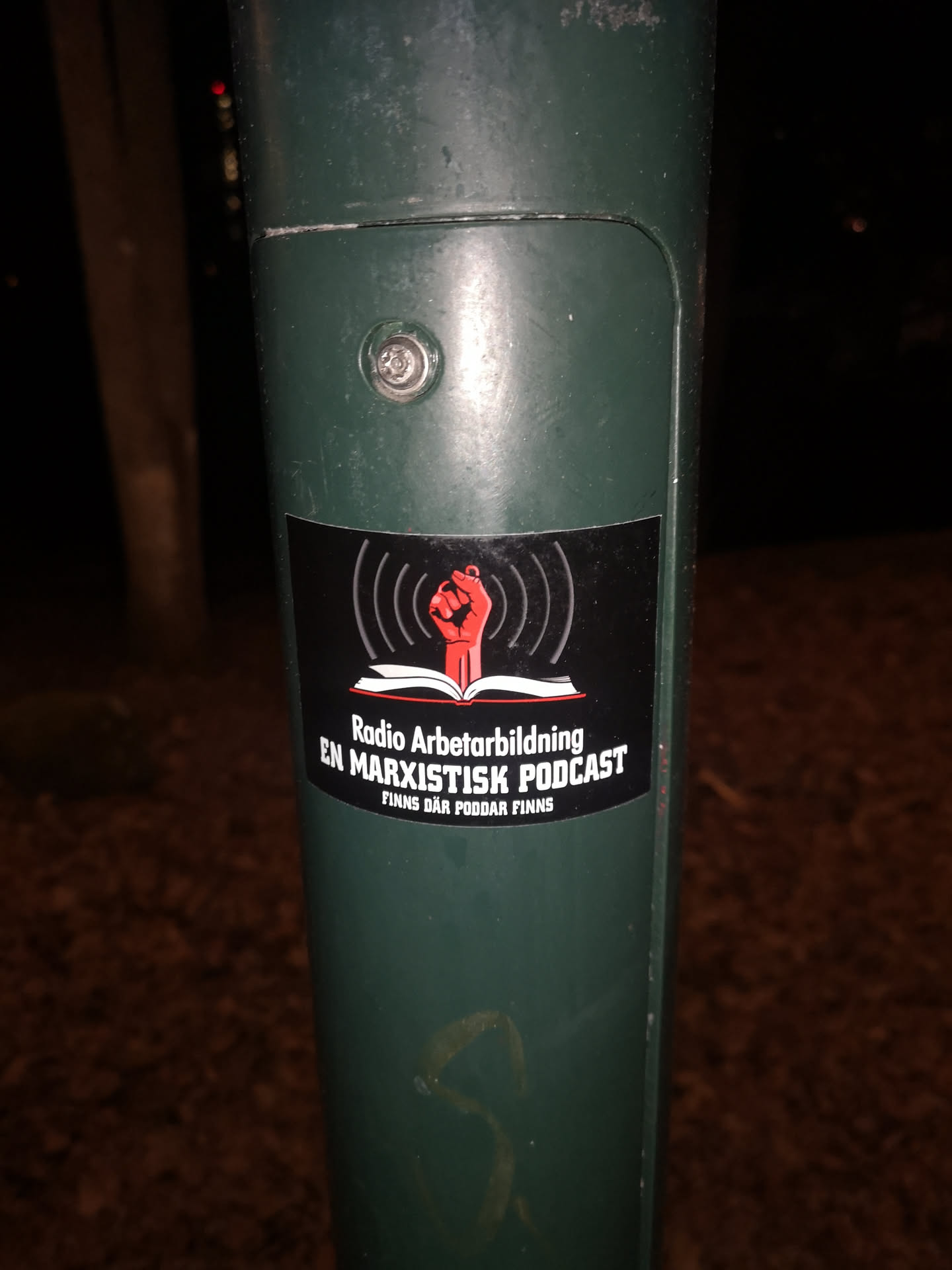 The sticker is a black rectangular design with slightly rounded corners, affixed to a dark green pole. It features a central graphic of a red, clenched raised fist emerging from an open white book, symbolizing knowledge and solidarity. Above the fist, white curved lines represent radio waves, indicating a broadcast. Below the graphic, the text 'Radio Arbetarbildning' is prominently displayed in white, followed by 'EN MARXISTISK PODCAST' in bold, all-caps white lettering. At the very bottom, in smaller white text, it reads 'FINNS DÄR PODDAR FINNS'. The overall aesthetic is stark and impactful, using red, white, and black.