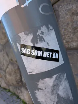 Street sticker A rectangular black sticker with bold white Swedish text reading 'SÄG SOM DET ÄR'. It is affixed over a torn, weathered white square sticker on a grey metal post.