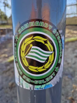 A round sticker featuring the Hammarby Stockholm logo. The logo includes a flag with green and white stripes, surrounded by a yellow wreath. The text "HAMMARBY STOCKHOLM" is written in a circular fashion around the logo, and the text "SM-GULDET" is partially visible at the bottom.