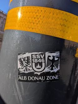 Street sticker The sticker is rectangular with a white border, featuring a monochromatic design. The central element is a shield divided into two halves. The left half contains a black eagle with two heads, stylized with sharp lines. The right half displays the text 'SSV 1846' in a bold, sans-serif font above a stylized mountain or crest shape, also in black and white. Below the shield, the text 'ALB DONAU ZONE' is prominently displayed in white capital letters against a black background with a distressed, brush-stroke effect. The overall aesthetic is bold and emblem-like.