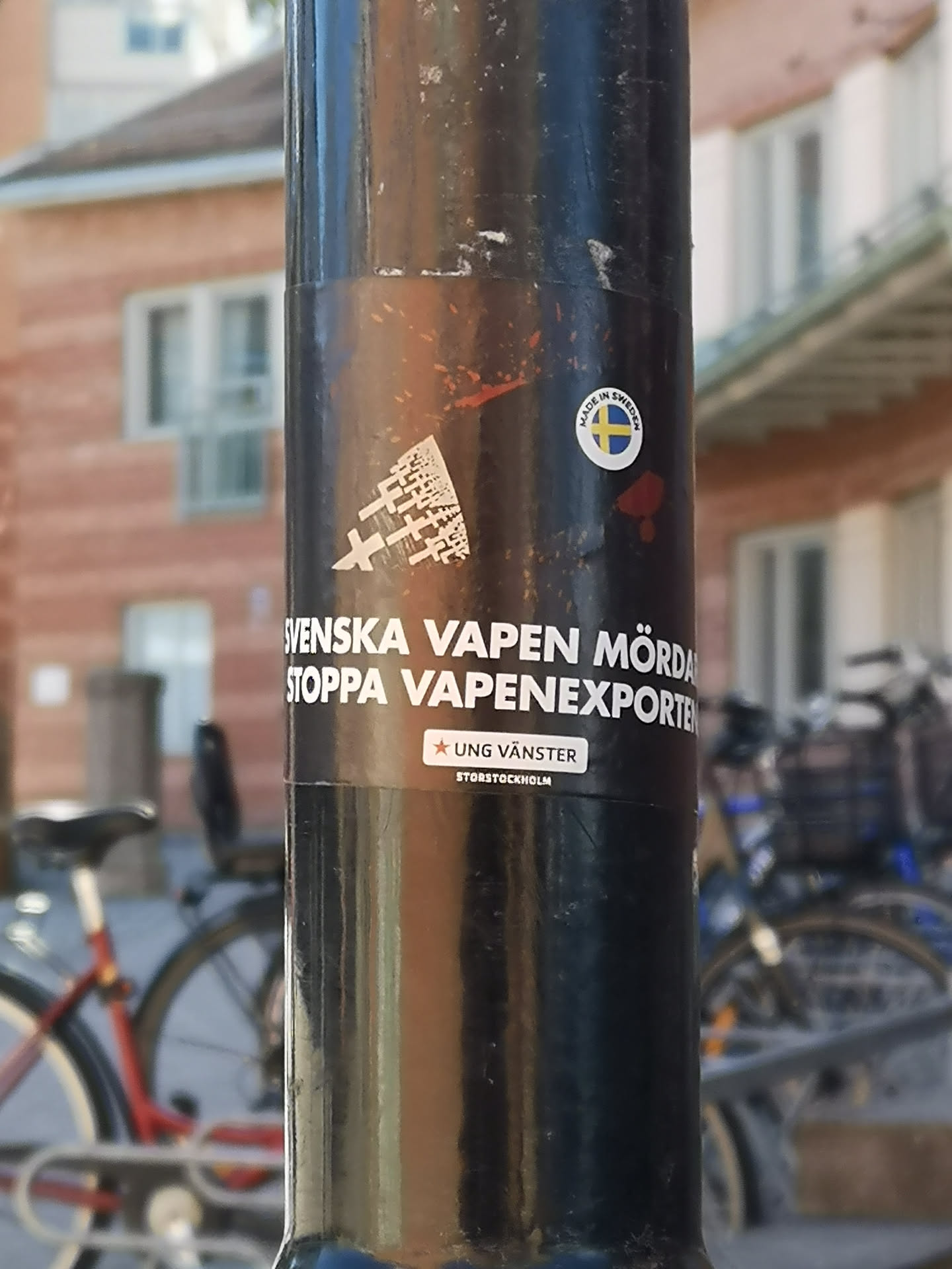 The central sticker is a dark, rectangular decal with white text and graphics, affixed to a black metal pole. At the top left, there's a white abstract graphic resembling fragmented 'X' shapes or a deconstructed weapon. Below this, the main message is prominently displayed in white, sans-serif capital letters: 'SVENSKA VAPEN MÖRDAR STOPPA VAPENEXPORTEN' (Swedish weapons kill, stop weapon exports). Further down, a smaller white rectangular box contains a red star icon followed by 'UNG VÄNSTER' (Young Left), and below that, 'STORSTOCKHOLM' (Greater Stockholm). Slightly above and to the right of the main sticker, there's a smaller circular sticker with a white border, featuring the text 'MADE IN SWEDEN' encircling a depiction of the Swedish flag. The sticker shows some minor wear and tear, consistent with being outdoors.