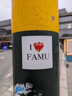 Street sticker The sticker is square-shaped with a white background. Centered on the sticker is a stylized red heart engulfed in flames, with yellow and orange hues at the base of the flames. Above the heart, the letter 'I' is printed in a dark, serif font. Below the heart, the word 'FAMU' is printed in a larger, dark, serif font, matching the style of the 'I'. The sticker appears to be made of paper and is affixed to a dark green pole.