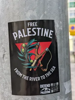 A sticker with a black background and white text that says "FREE PALESTINE" in large letters. Below the text is an image of an olive branch and a slingshot in a red triangle. The text "FROM THE RIVER TO THE SEA" is at the bottom of the sticker. At the very bottom, there is a logo that says "DEFEND PALESTINE" with a picture of an assault rifle. There is also a QR code on the sticker.