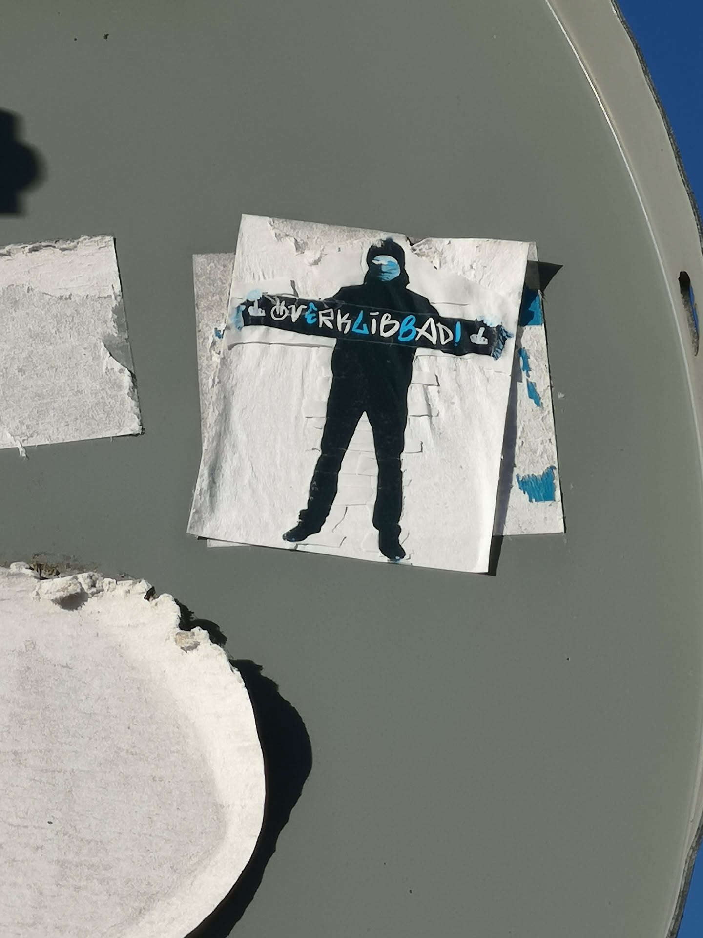 A monochrome sticker featuring a stylized, dark silhouette of a person standing, possibly wearing a hoodie or beanie. The figure's face is partially obscured by a light blue face mask. They hold a long, horizontal banner or scarf across their chest, which is dark with light blue text that reads 'OVERKLIBBAD!' and a small symbol resembling a fist or hand gesture at the right end. The sticker itself is white, showing signs of wear and peeling at the edges, and is affixed to a grey, curved surface.