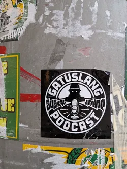 Street sticker A square black sticker featuring a circular white emblem. Inside the circle, there is a silhouette of a person wearing a hat with glowing white eyes, positioned behind a classic vintage microphone with radiating sound lines. The text 'GATUSLANG' is arched across the top, 'PODCAST' is arched across the bottom, and the words 'SVENSK' and 'HIPHOP' are written in a stylized font on either side of the microphone.