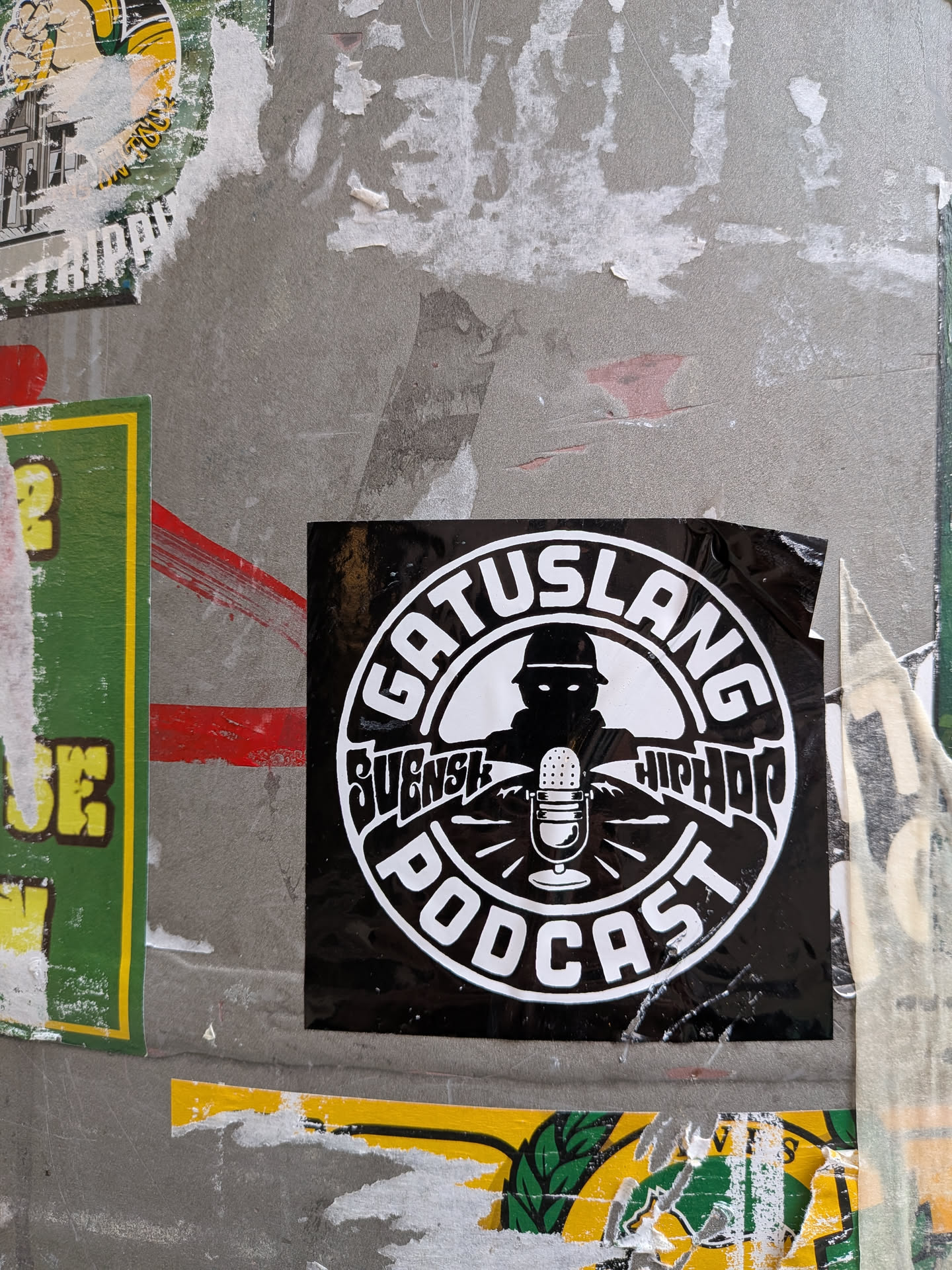 A square black sticker featuring a circular white emblem. Inside the circle, there is a silhouette of a person wearing a hat with glowing white eyes, positioned behind a classic vintage microphone with radiating sound lines. The text 'GATUSLANG' is arched across the top, 'PODCAST' is arched across the bottom, and the words 'SVENSK' and 'HIPHOP' are written in a stylized font on either side of the microphone.