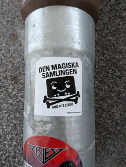Street sticker A rectangular white sticker with a black and white design. At the top, in bold uppercase sans-serif font, is the text "DEN MAGISKA SAMLINGEN". Below this, a stylized, blocky black character with a wide smile, two small dot eyes, and two small curved lines above its eyes is depicted. This character's head replaces the traditional skull in a skull and crossbones motif, with two thick, stylized black crossbones beneath it. Below the character and crossbones, in smaller bold uppercase sans-serif font, is the text "AND IT'S LEGAL". At the very bottom, in tiny text, is "DENMAGISKASAMLINGEN.SE". The sticker has slightly worn edges, consistent with being adhered to a metal pole.