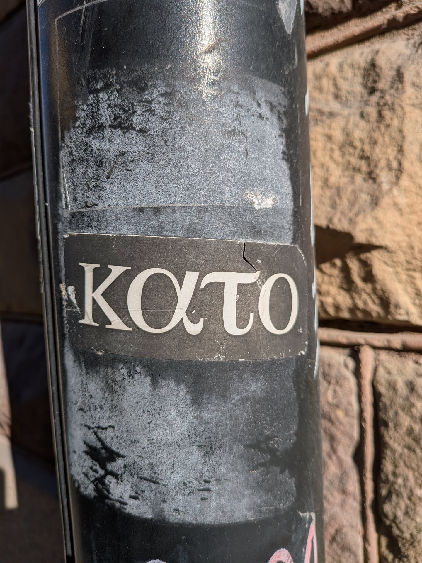 A rectangular sticker featuring the word 'KATO' written in white or light grey serif Greek letters against a dark grey or black background. The sticker shows signs of wear, including a visible crack running through the letter 'τ' (tau) and some peeling around the edges. The overall appearance is simple and text-focused, with a clear contrast between the lettering and the background.