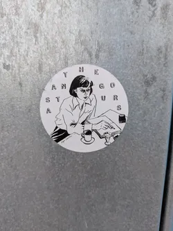 The sticker is circular and features a black and white illustration of a young woman with short hair, wearing a collared shirt and striped pants. She is seated at a table, with her head tilted down as she reads a book or document. Her left hand rests on the page, holding a pen. To her right, there is a cup of coffee on a saucer and a small glass. The text 'THE GURUS' is arranged in a circular pattern above her, with some letters incomplete. Below her, the text 'Kajsa Danielsson' is written in a cursive font.