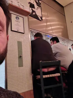 Street sticker This image shows the back of a person sitting on a bar stool.  The focus is primarily on the person's rear and the stool itself. The background includes other bar patrons and a tiled wall.