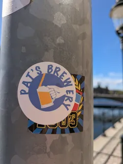A round sticker with 'Pat's Brewery' written around the edge in blue with a drawing of a hand holding a beer. Below it, there is a 2003 sticker with a black and blue striped background.
