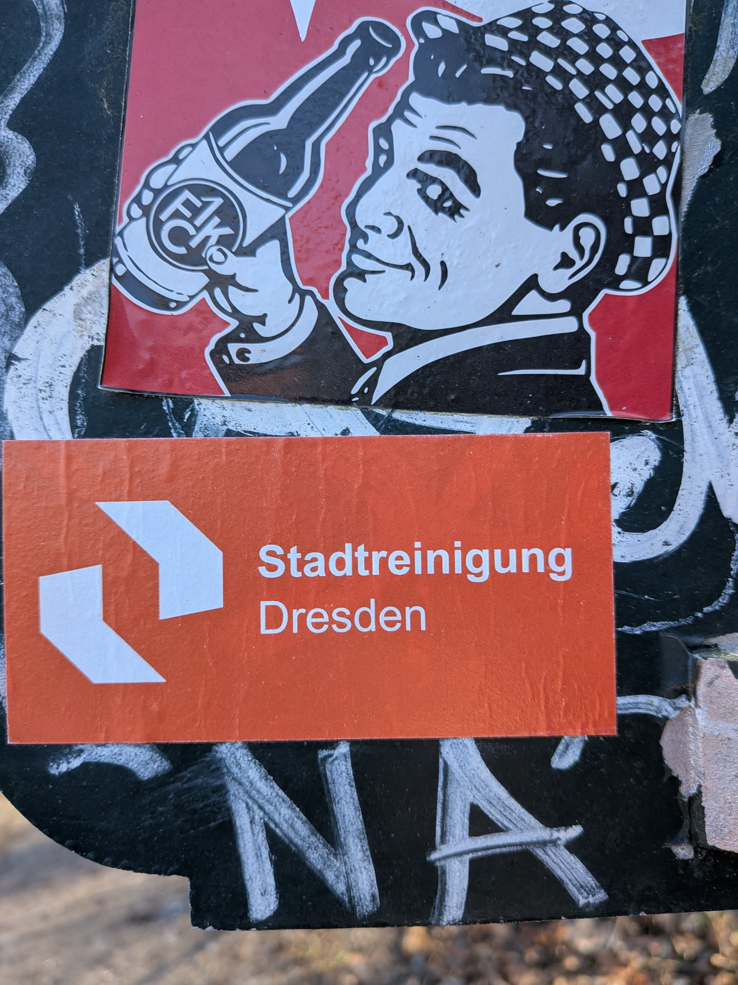 A rectangular orange sticker featuring two white, angular arrow-like shapes on the left side, pointing towards the right. To their right, in white sans-serif text, are the words 'Stadtreinigung' on the upper line and 'Dresden' on the lower line. The sticker has a slightly textured appearance and minor creases, suggesting it is a physical adhesive label.