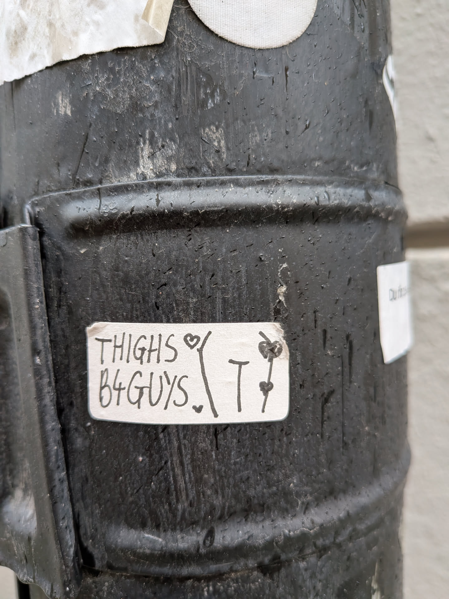 A rectangular, off-white sticker with slightly rounded corners, adhered to a dark, textured metal surface. The sticker features handwritten black text that reads 'THIGHS B4 GUYS'. A small black heart is drawn above the 'S' in 'THIGHS' and another below the 'S' in 'GUYS'. To the right of the main text, there is a larger, stylized black 'T' with two small black hearts drawn on it, one near the top and one near the bottom. The sticker appears slightly worn and has a homemade aesthetic.