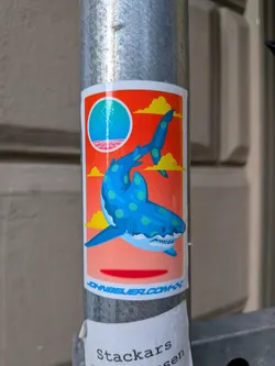 Street sticker The sticker features a stylized blue shark with light green polka dots, swimming against a vibrant orange background. Above the shark, a circular motif depicts a blue orb with lightning striking from it, set against a pink horizon and stylized yellow clouds. The overall aesthetic has a retro-futuristic feel. The shark has its mouth open, revealing sharp white teeth. The bottom edge of the sticker has a white border with blue text.