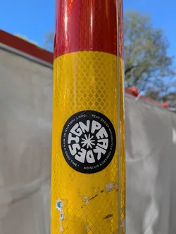 Street sticker A circular black sticker with white text and a central graphic, adhered to a yellow reflective pole. The central graphic is a stylized white star or flower-like emblem. Around the top edge, in an arc, is the text "PEAK DESIGN". Around the bottom edge, also in an arc, is "MOTORCYCLE RESEARCH DIVISION". On the left side, in a larger arc, is the quote "DON'T THREATEN ME WITH A GOOD TIME." The text is clear and sans-serif, creating a bold, minimalist design.