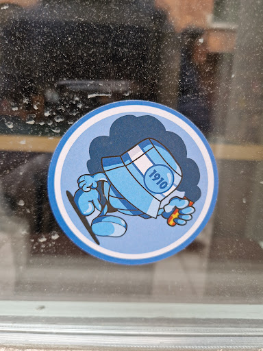 A round sticker featuring a cartoon character. The character is light blue, wearing a bucket hat with "1910" written on it and appears to be holding a small orange object. It's set against a darker blue cloud-like background. The style is simple and slightly reminiscent of classic cartoon characters.