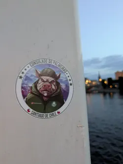 Street sticker The sticker is circular with a white border. At its center is an illustration of a pig wearing a green baseball cap with a 'P' logo and a green hooded jacket. The pig has a stern expression. Behind the pig is a mountainous landscape in shades of purple and blue, with a few white peaks. Around the top edge of the sticker, in a semi-circle, is the text "CONSULADO DO PALMEIRAS" surrounded by small stars. Along the bottom edge, also in a semi-circle, is the text "SANTIAGO DE CHILE", with small red chili peppers on either side. There is also a small Instagram handle visible on the pig's jacket.