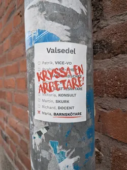 Street sticker A white rectangular sticker, designed to resemble a ballot paper, is affixed to a grey metal pole. It features a list of names with associated professional titles in black text, headed by 'Valsedel' (Ballot paper). Prominently overlaid in bold, red, handwritten script are the words 'KRYSSA EN ARBETARE!' (Cross a worker!). A red 'X' marks the checkbox next to 'Maria, BARNSKÖTARE' (childcare worker). The sticker exhibits signs of outdoor exposure, including slightly curled edges and minor wear.