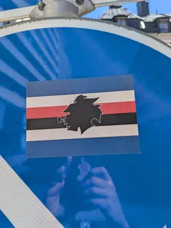 Street sticker The sticker is rectangular, displaying the distinctive flag design of U.C. Sampdoria. The flag consists of five horizontal stripes: a wide blue stripe at the top, followed by a thinner white stripe, a red stripe, a black stripe, another white stripe, and a wide blue stripe at the bottom. Superimposed and centered on these stripes is a black silhouette of "Baciccia," a traditional Genoese character depicted in profile, wearing a cap and smoking a pipe. The sticker appears slightly worn, with minor creases or imperfections, particularly visible at the corners. It is affixed to a reflective blue surface, which has a grid-like pattern.