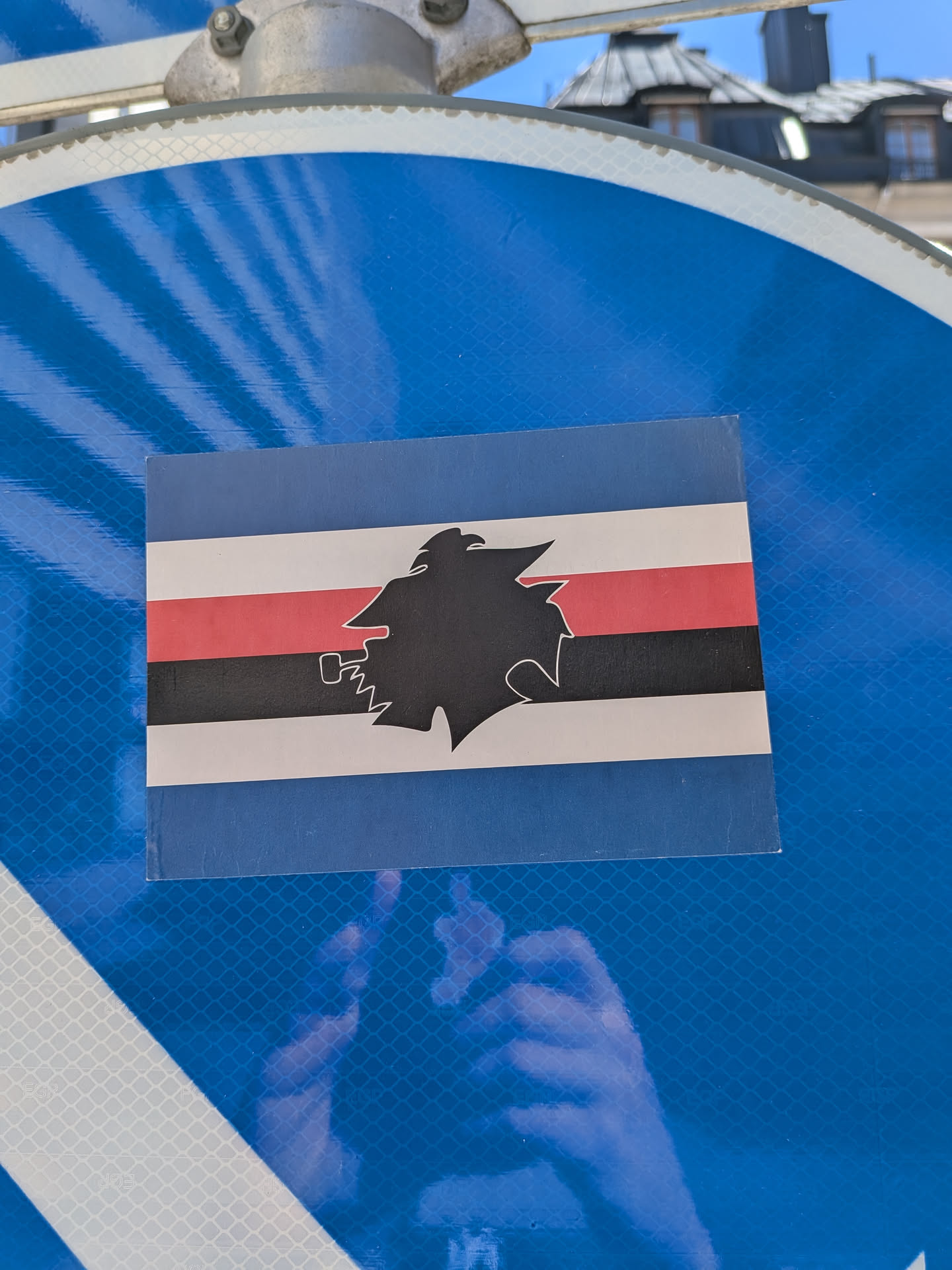 The sticker is rectangular, displaying the distinctive flag design of U.C. Sampdoria. The flag consists of five horizontal stripes: a wide blue stripe at the top, followed by a thinner white stripe, a red stripe, a black stripe, another white stripe, and a wide blue stripe at the bottom. Superimposed and centered on these stripes is a black silhouette of "Baciccia," a traditional Genoese character depicted in profile, wearing a cap and smoking a pipe. The sticker appears slightly worn, with minor creases or imperfections, particularly visible at the corners. It is affixed to a reflective blue surface, which has a grid-like pattern.