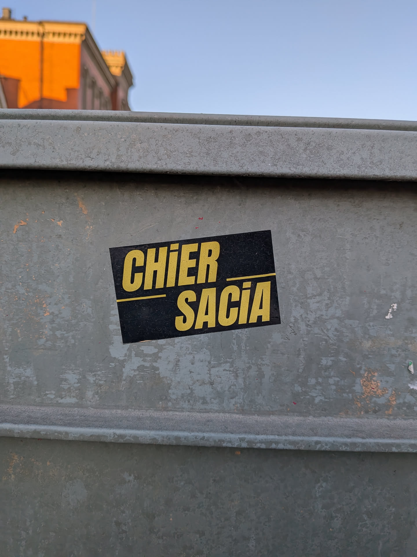 The sticker is a rectangular, horizontally oriented design with a solid black background. It features two lines of prominent, bold, sans-serif text in a bright yellow color. The top line reads 'CHIER' and the bottom line reads 'SACIA'. A thin, horizontal yellow line separates the two words. The sticker is affixed to a textured, weathered grey-green metal surface, possibly a utility box or dumpster, showing some minor wear and slight peeling at its edges.