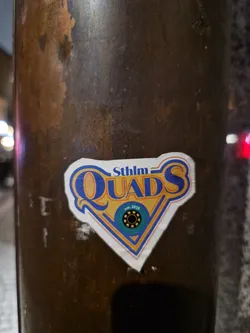 Street sticker A sticker featuring the logo for Stockholm Quads, a rollerblading group. The logo is a stylized diamond shape with the text 'Sthlm QUADS' in blue and yellow, and a smaller circular logo inside. The sticker is affixed to a dark brown metal pole.