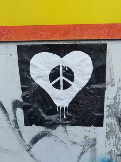 A black and white sticker on a wall shows a white heart shape with a peace symbol inside.  The heart appears to be dripping. The sticker is affixed to a wall with some existing graffiti.