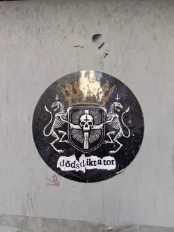 Street sticker The sticker is a round, black and white design with a golden crown at the top. Below the crown is a heraldic shield with a cross and a skull. Two skeletal lions flank the shield, each holding a sword. At the bottom, the word "dödsdiktator" is written in a rough, stylized font.