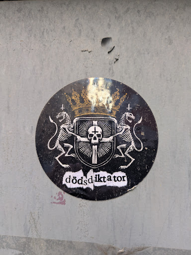Street sticker The sticker is a round, black and white design with a golden crown at the top. Below the crown is a heraldic shield with a cross and a skull. Two skeletal lions flank the shield, each holding a sword. At the bottom, the word "dödsdiktator" is written in a rough, stylized font.