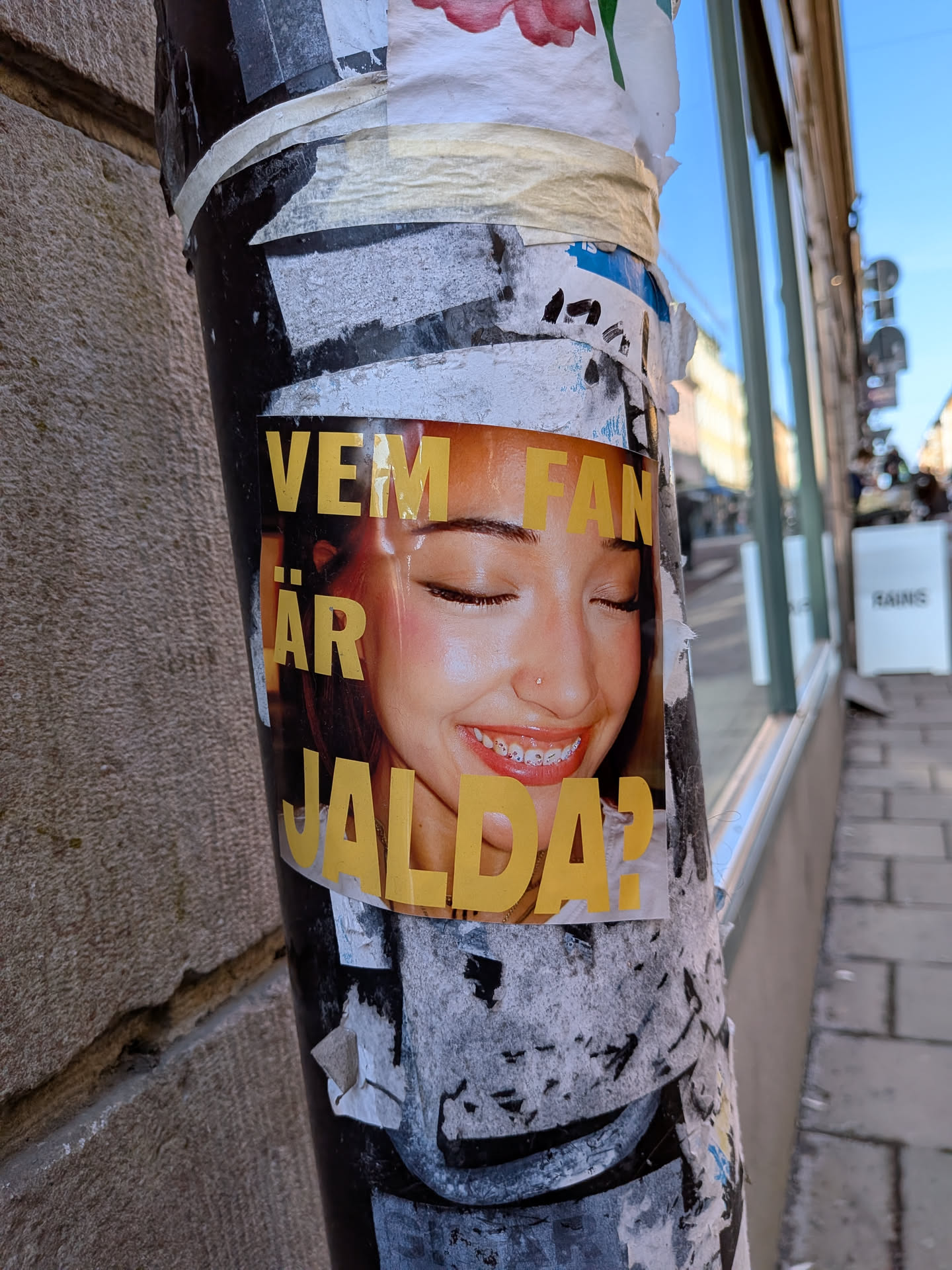 A rectangular sticker, vertically oriented, featuring a close-up portrait of a young woman's face. She has her eyes closed, a gentle smile revealing braces on her teeth, and a small stud nose piercing. Her skin has warm, radiant tones. Overlaid on the image is bold, yellow, sans-serif text that reads 'VEM FAN ÄR JALDA?'. The background of the portrait is a soft, blurred orange-brown. The sticker is adhered to a heavily worn and layered black pole, surrounded by other torn paper fragments.