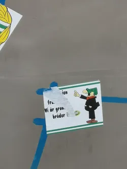 Street sticker A rectangular sticker with a white background and a green and white striped border along the top and bottom edges. On the right side, a stylized cartoon character is depicted, featuring a light-colored face, a black suit, an orange/red scarf, and a distinctive green plaid flat cap. The character is pointing with their right hand towards the left. The left side of the sticker is partially obscured by white adhesive residue and some tearing. Visible text on the left reads 'Vi är grön. bröder'. Below this text, there's a small circular green logo with a wavy line inside.