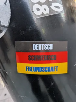 Street sticker The sticker is rectangular with rounded corners and features three horizontal bands of color. The top band is black with white text that reads 'DEUTSCH'. The middle band is red with black text that reads 'SCHWEDISCH'. The bottom band is yellow with blue text that reads 'FREUNDSCHAFT'. The overall design evokes the German flag.