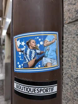 Street sticker Rectangular sticker showing a female footballer in a blue-and-white striped kit kicking a player in light blue. Set against a blue background with white stars and a yellow border.
