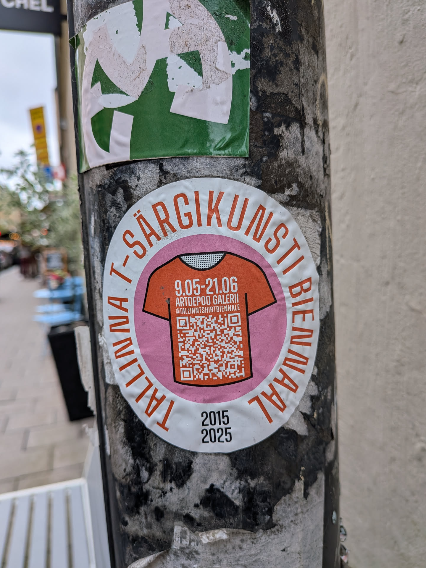 An oval-shaped sticker with a white border featuring the orange text 'TALLINNA T-SÄRGIKUNSTI BIENNAAL' curved around the top and sides, and the years '2015' and '2025' printed in black at the bottom. The center contains a pink circle enclosing an orange T-shirt graphic. Inside the T-shirt illustration, white text lists the dates '9.05-21.06', the venue 'ARTDEPOO GALERII', and the Instagram handle '@TALLINNTSHIRTBIENNALE', above a large QR code.