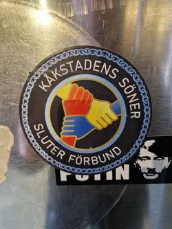 Street sticker Round sticker with a black background and blue and white chain-like border. It features three hands clasped together in the center, colored red, yellow and blue. The text around the image says Käkstadens Söner Sluter Förbund.