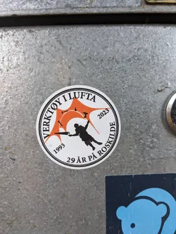 Street sticker A round sticker commemorating 29 years of Verktoy i Lufta, an event that has taken place in Roskilde since 1993. The sticker features an illustration of a person working with tools against an orange background, with the years 1993 and 2023 displayed on either side of the image. The text around the circular border states "Verktoy i Lufta" and "29 år på Roskilde".