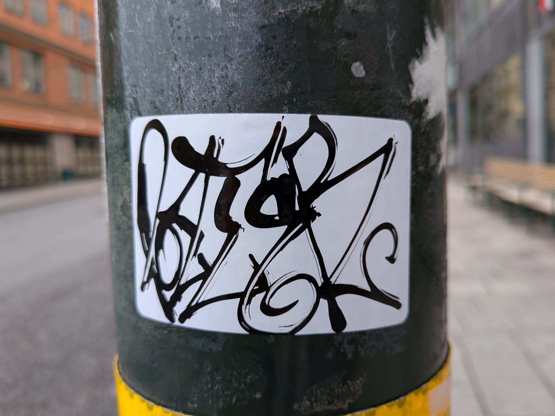 The sticker is rectangular with rounded corners and features a stylized graffiti tag in bold black ink on a clean white background. The tag appears to be an abstract representation of letters, characterized by sharp angles, fluid curves, and drips. The overall impression is energetic and artistic.