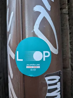 Street sticker A circular teal-colored sticker prominently featuring the brand name 'L O P' in large white capital letters. The 'O' is represented by a solid white circle, with several horizontal white lines extending from the 'L' towards it, creating a dynamic visual effect. Below the main logo, the flavor 'JALAPEÑO LIME' is written in smaller white capital letters. Further down, the word 'STRONG' is displayed in white, accompanied by a visual strength indicator consisting of three solid red circles and two outlined white circles, suggesting a high potency. A small white oval contains the text '18+' and 'HOT!', indicating an adult product with a spicy profile. The sticker is affixed to a textured, dark brown pole or pipe, with remnants of white graffiti visible around its edges.