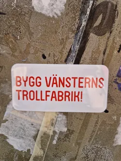 Street sticker A white sticker with red text on a grunge background. The text says "BYGG VÄNSTERNS TROLLFABRIK!" which translates to "Build the Left's troll factory!"