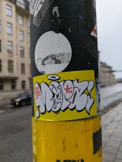A yellow sticker with graffiti art is attached to a pole. The sticker features stylized lettering and a cartoon character with an angry expression. The character has a halo above its head and a red star next to it. The style is reminiscent of street art or bombing.