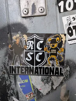 Street sticker The sticker features two cartoon characters flanking a shield with 'RC SC' in large white letters. On the left is a cowboy wearing a tan hat, a black hoodie with a white logo (partially obscured), and blue jeans. He holds a lit flare in his right hand. On the right is a cheetah-like character wearing a black bandana, a black t-shirt with 'ULTRAS CHARLEROI' written in white, and white sneakers with black stripes. This character also holds a lit flare. Both characters have smoke emanating from their flares. The word 'INTERNATIONAL' is prominently displayed in large, stylized white block letters below the shield. The background of the sticker is dark with subtle textures, and the entire design appears to be printed on a rectangular sticker.