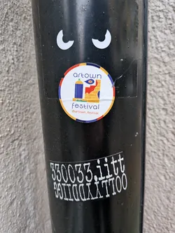 Street sticker A circular sticker featuring a colorful illustration. It has an outer orange-red ring, followed by a blue ring, and a white central background. The main graphic depicts a blue spray paint can with a green nozzle on the left, next to a red and yellow brick wall on the right. An eye symbol is visible on the yellow section of the wall. Above the illustration, 'artown' is written in purple, and below it, 'festival' is in red, followed by '@artown_festival' in purple.