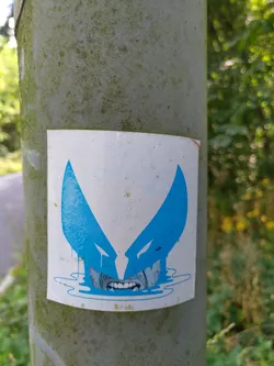 A sticker featuring a stylized blue Wolverine face, seemingly melting or dripping, affixed to a weathered pole. The art style is simple yet striking.